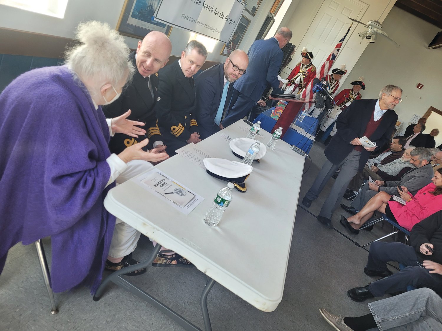 THE SEARCHERS: From left to right, Dr. Kathy Abbass, British Naval Commanders Steven White and Simon Rogers, Dr. Peter Abbott, state Rep. Joseph M. McNamara, the Pawtuxet Rangers, and Gaspee Days Committee board member Roger Hudson prepare for Tuesday morning’s announcement at the Aspray Boathouse in Pawtuxet Village. (Beacon photo by Rory Schuler)